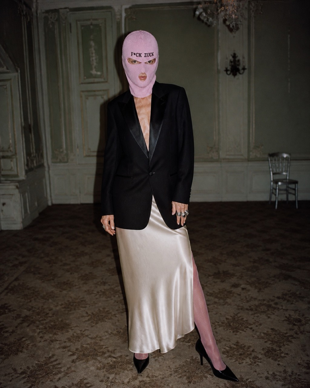 Model in the blush Zucks Blindspot balaclava wearing a tuxedo jacket and satin skirt in a faded ballroom.